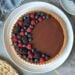 A dark chocolate tart with fresh berries arranged in a half crescent moon on top of it sit on a large ceramic serving plate.