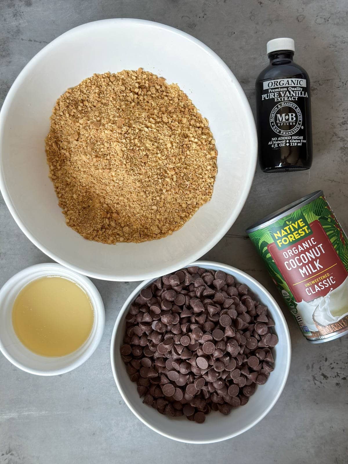A large white ceramic bowl contains graham cracker crumbs, while a can of coconut milk, chocolate chips in a small white bowl, melted coconut oil, and a bottle of vanilla extract surround the larger bowl.