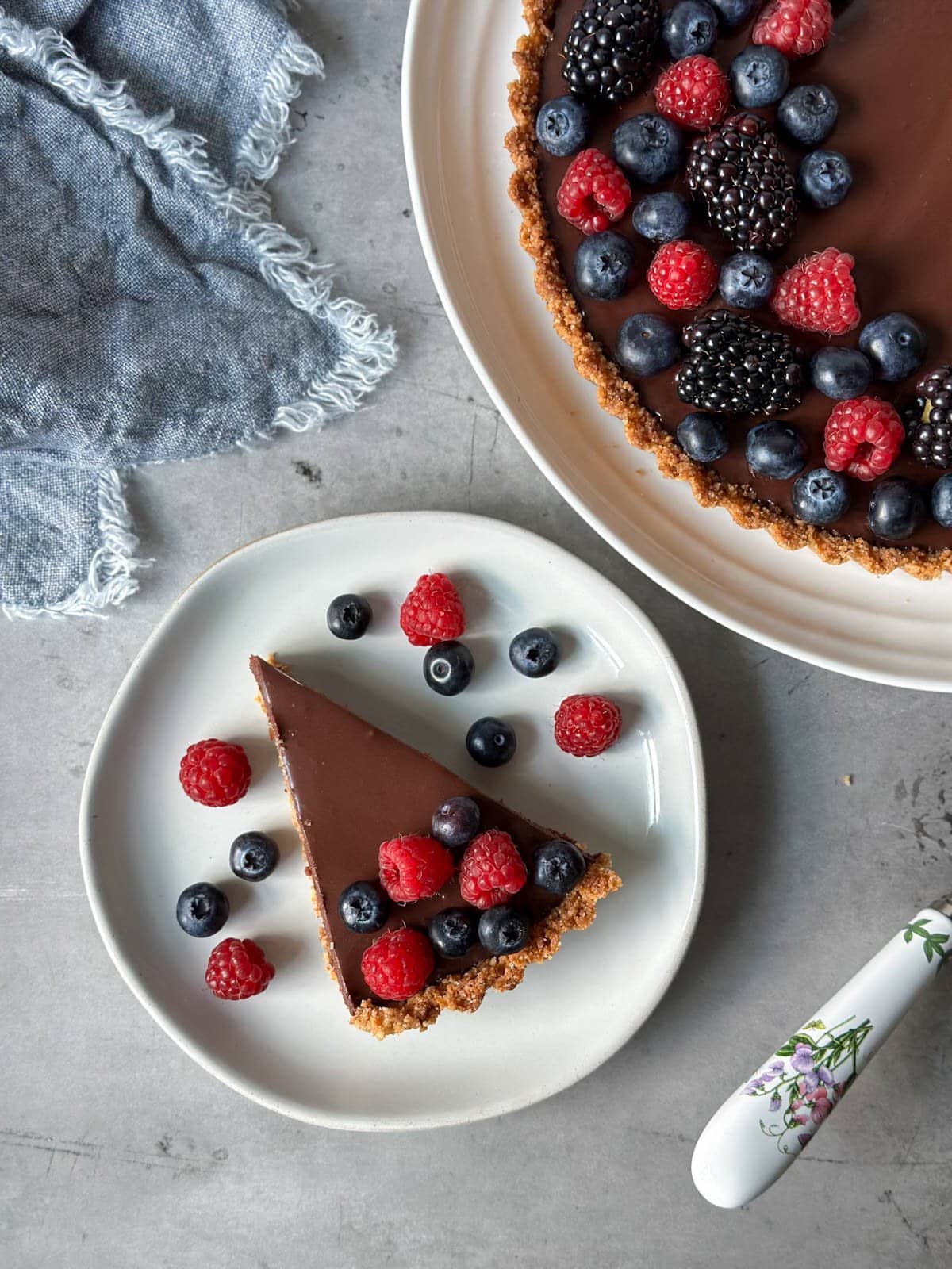 A small ceramic plate has a slice of a chocolate tart, it is covered in fresh raspberries and blueberries. Nearby is a large platter that contains the rest of the chocolate tart.