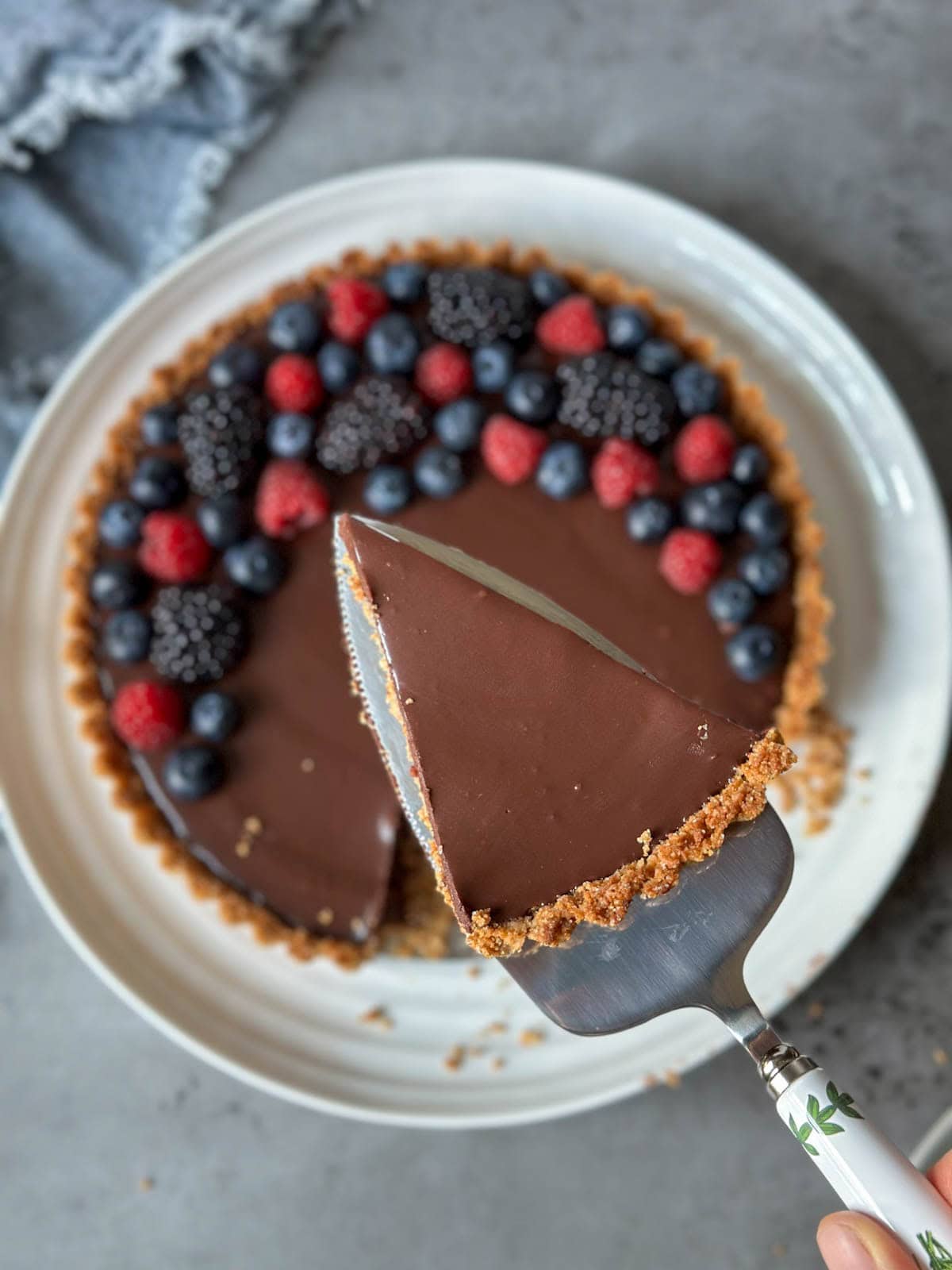 A cake serving utensil is held with a slice of chocolate tart on top of it. Below is the remainder of the tart on a large serving platter.