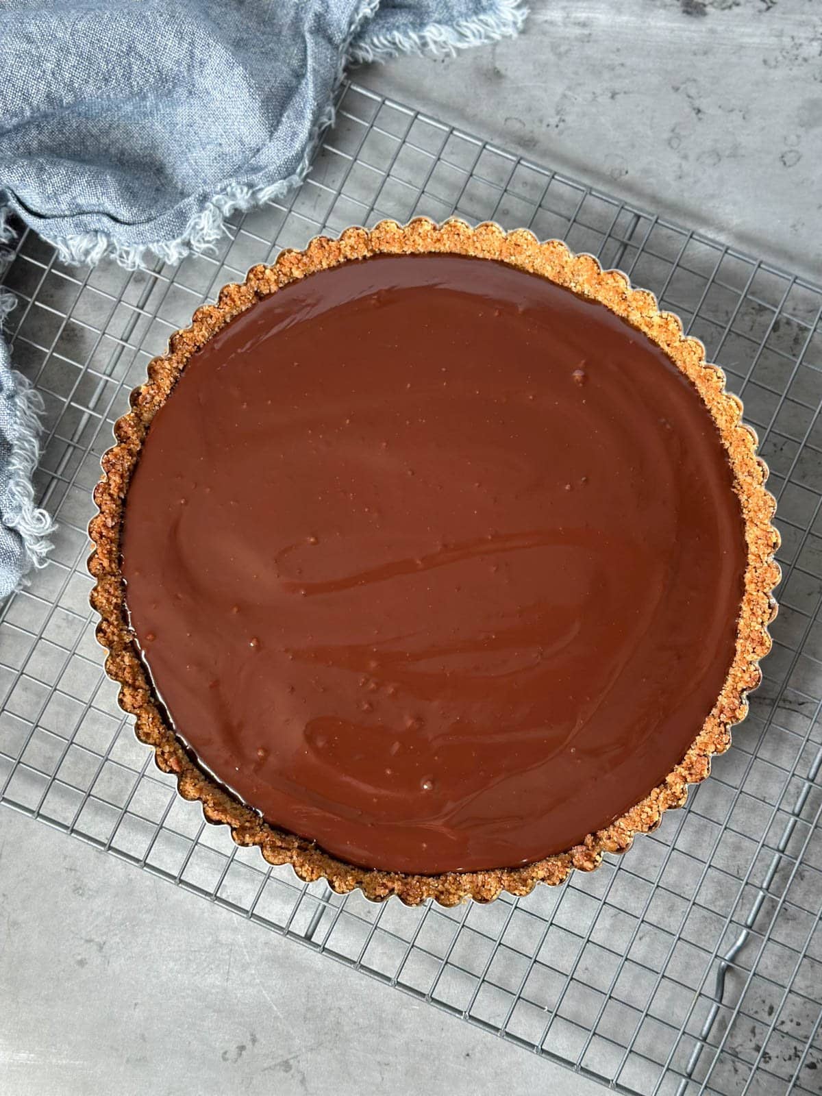 A chocolate tart with graham cracker crust sits on a wire cooling rack as the chocolate starts to set.