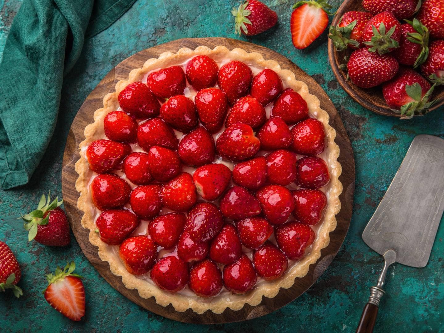 A strawberry pie is covered with fresh strawberries while additional fresh strawberries and a serving utensil surround the pie.