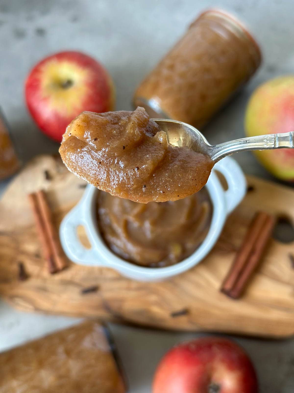 A silver spoon has a dollop of apple butter resting in it. Below is a ramekin of apple butter, cinnamon sticks, and whole apples.
