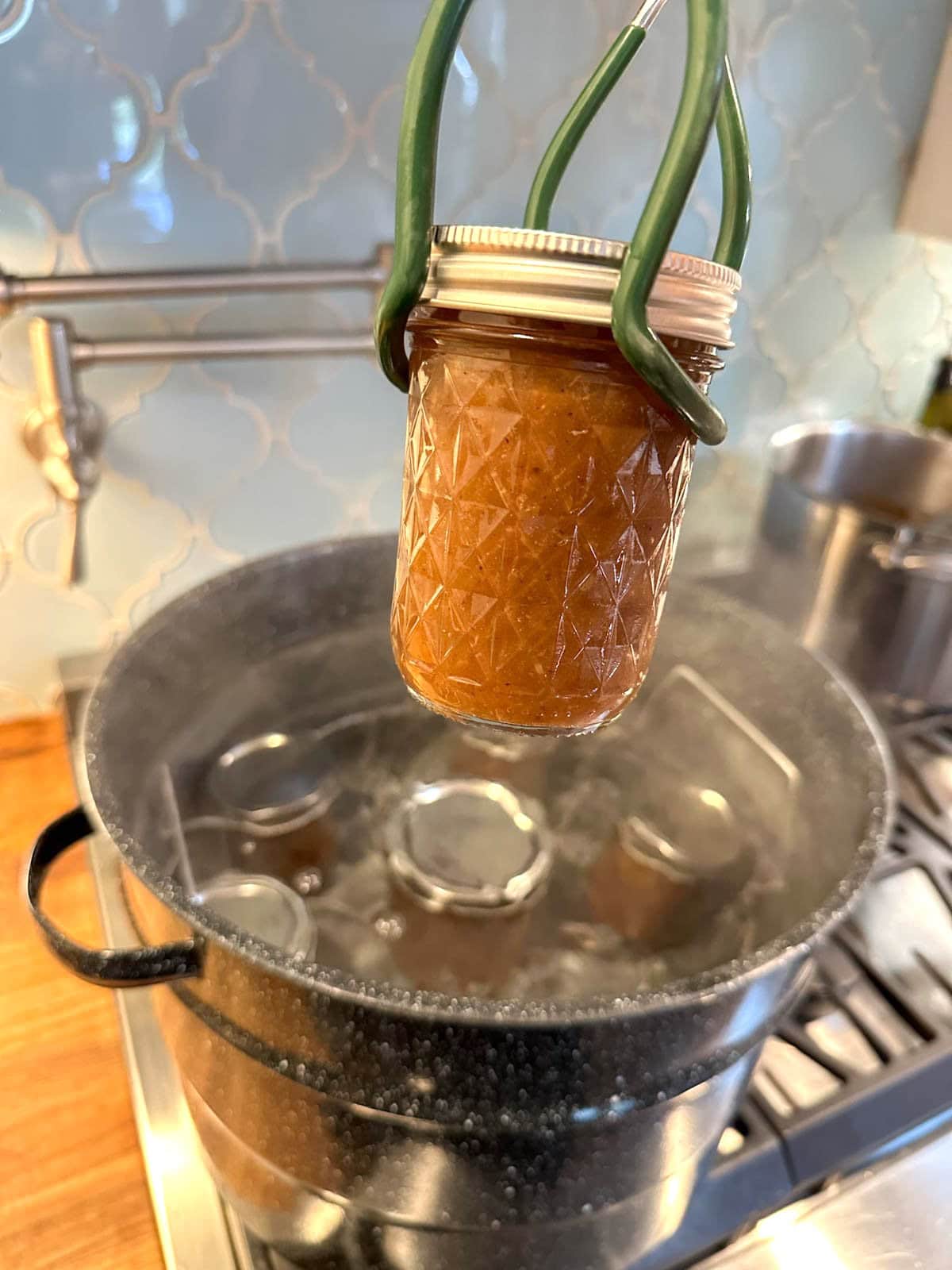 A jar lifter is holding a half pint jar, beyond is a canning pot filled with more half pint jars.