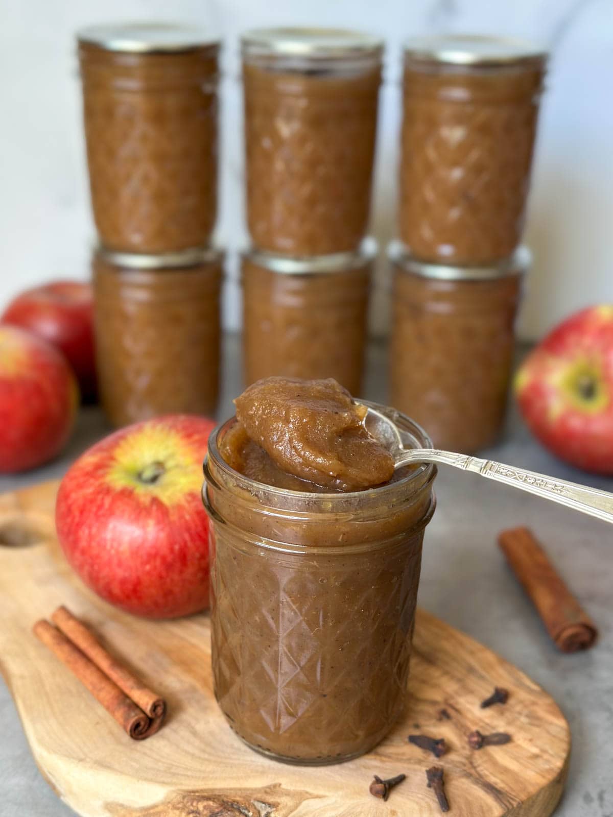 A half pint jar has a silver spoon resting on the top of it with a dollop of apple butter on it. Beyond are whole apples, cinnamon sticks, and more jars of apple butter.