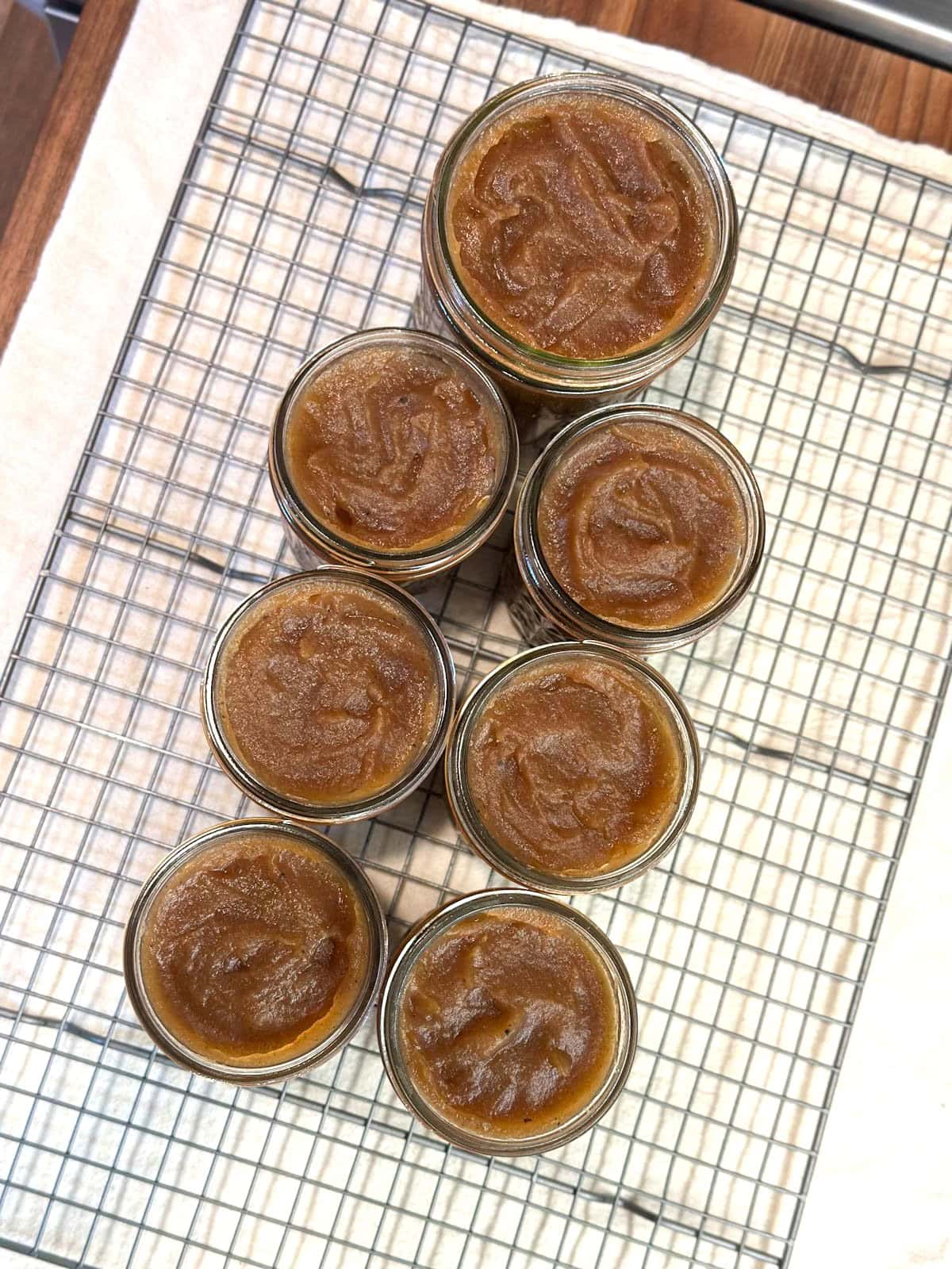 Six half pint jars and one pint jar sit on a wire mesh cooling rack full of apple butter.