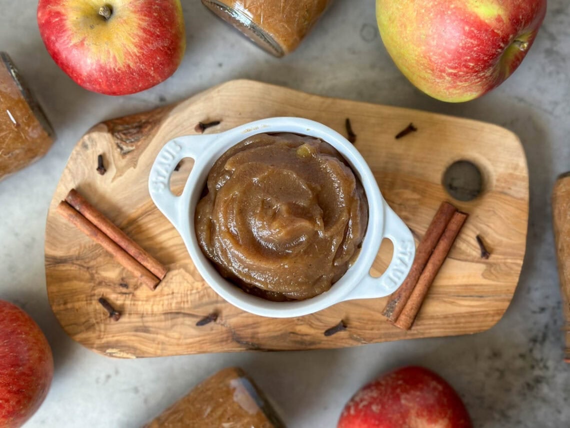 A white ramekin with handles is full of apple butter. Surrounding the ramekin are whole cinnamon stick, clove, and whole apples.
