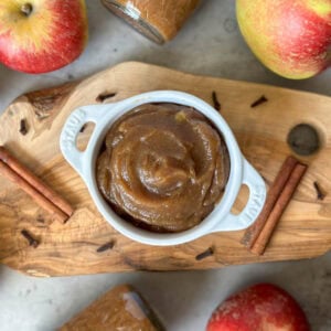 A white ramekin with handles is full of apple butter. Surrounding the ramekin are whole cinnamon stick, clove, and whole apples.
