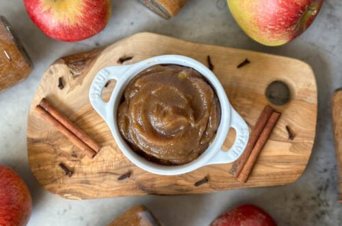 A white ramekin with handles is full of apple butter. Surrounding the ramekin are whole cinnamon stick, clove, and whole apples.