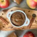 A white ramekin with handles is full of apple butter. Surrounding the ramekin are whole cinnamon stick, clove, and whole apples.