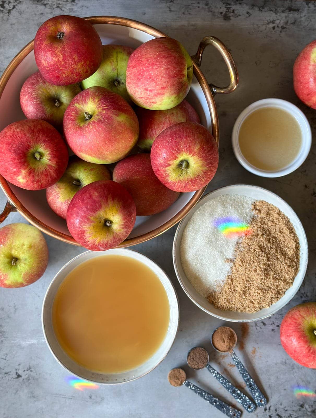 A large bowl contains apples mounded tall, a bowl of white and brown sugar, a bowl of apple cider, a small bowl of lemon juice along with cinnamon, clover, and allspice spices in measuring spoons. A few whole apples are scattered around the area.