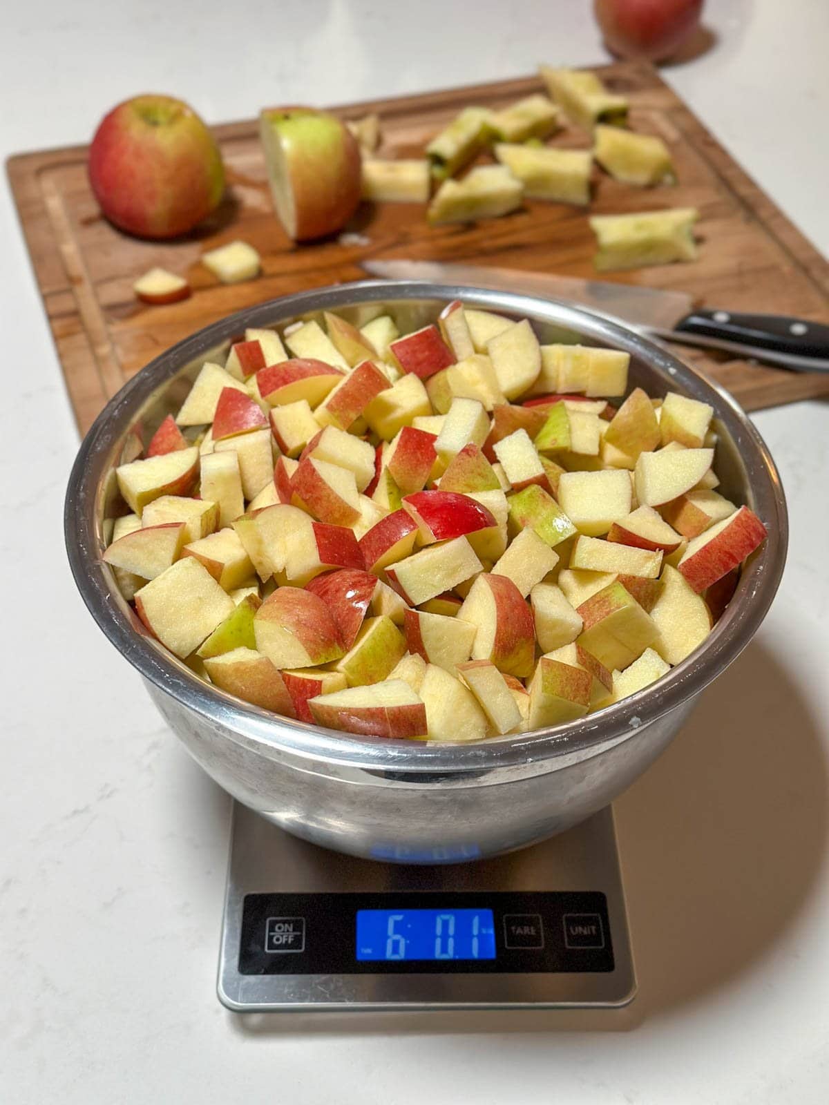 A large stainless steel bowl sits on a digital scale and is full of apple pieces that have been cut into one inch pieces.