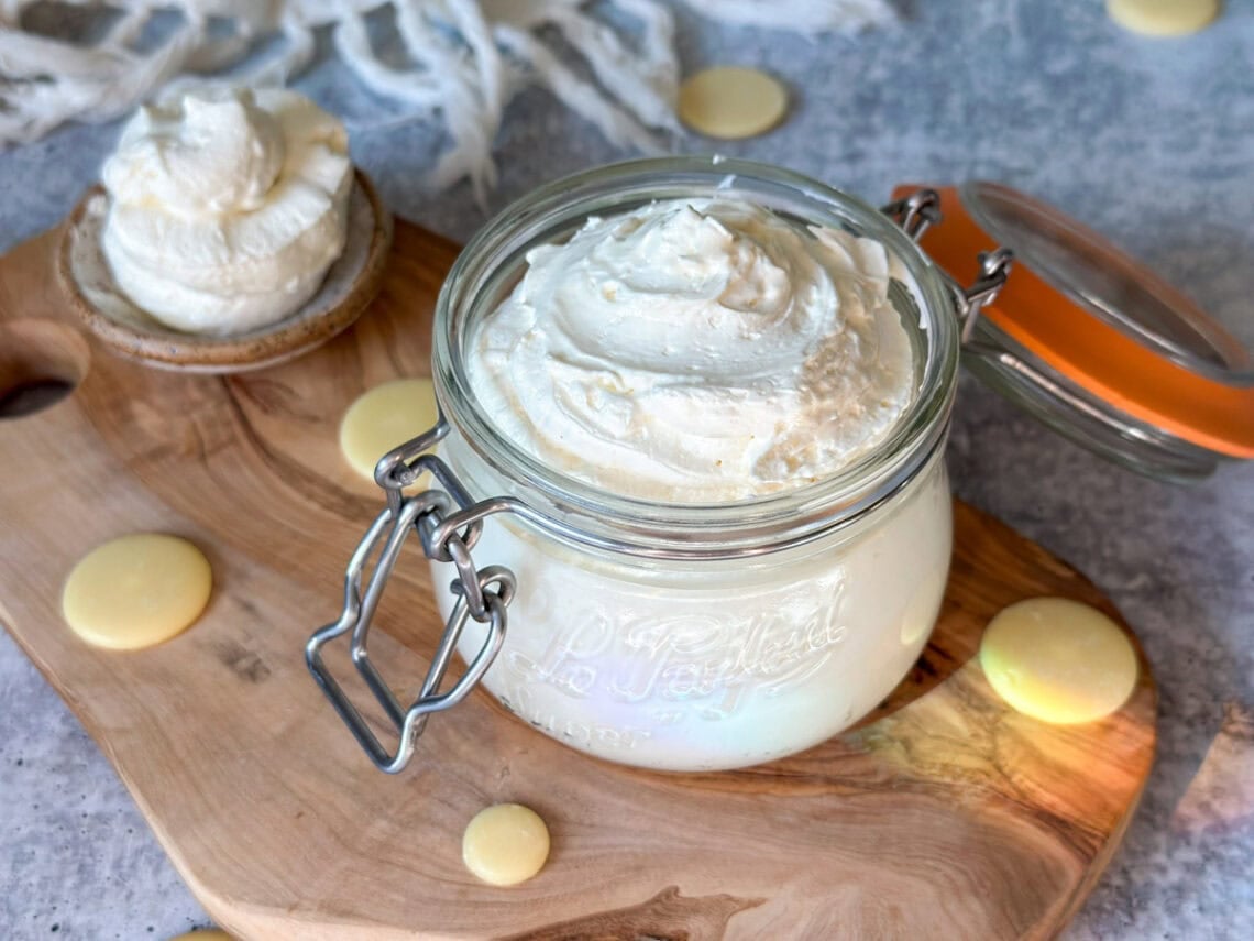 A close up of a Le Parfait 12-ounce glass jar with a flip-top lid full of fluffy white whipped body butter, swirled on top. There are also cocoa butter wafters laying around the base of the jar, and a large spoon full of whipped body butter laying off to the side