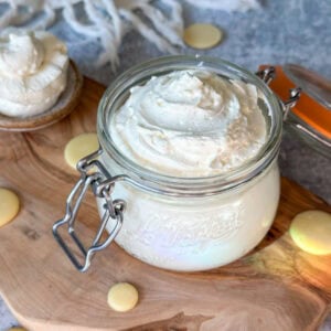 A close up of a Le Parfait 12-ounce glass jar with a flip-top lid full of fluffy white whipped body butter, swirled on top. There are also cocoa butter wafters laying around the base of the jar, and a large spoon full of whipped body butter laying off to the side