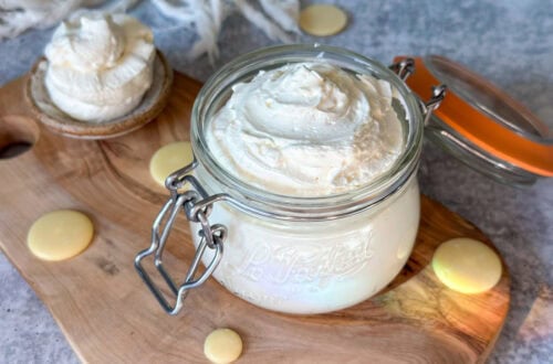A close up of a Le Parfait 12-ounce glass jar with a flip-top lid full of fluffy white whipped body butter, swirled on top. There are also cocoa butter wafters laying around the base of the jar, and a large spoon full of whipped body butter laying off to the side