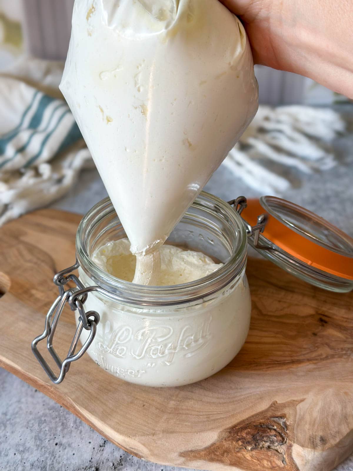 A hand is holding a ziplock bag that has been filled with whipped body butter, with a hole cut in the corner to squeeze the body butter into a glass jar below (like a piping bag)