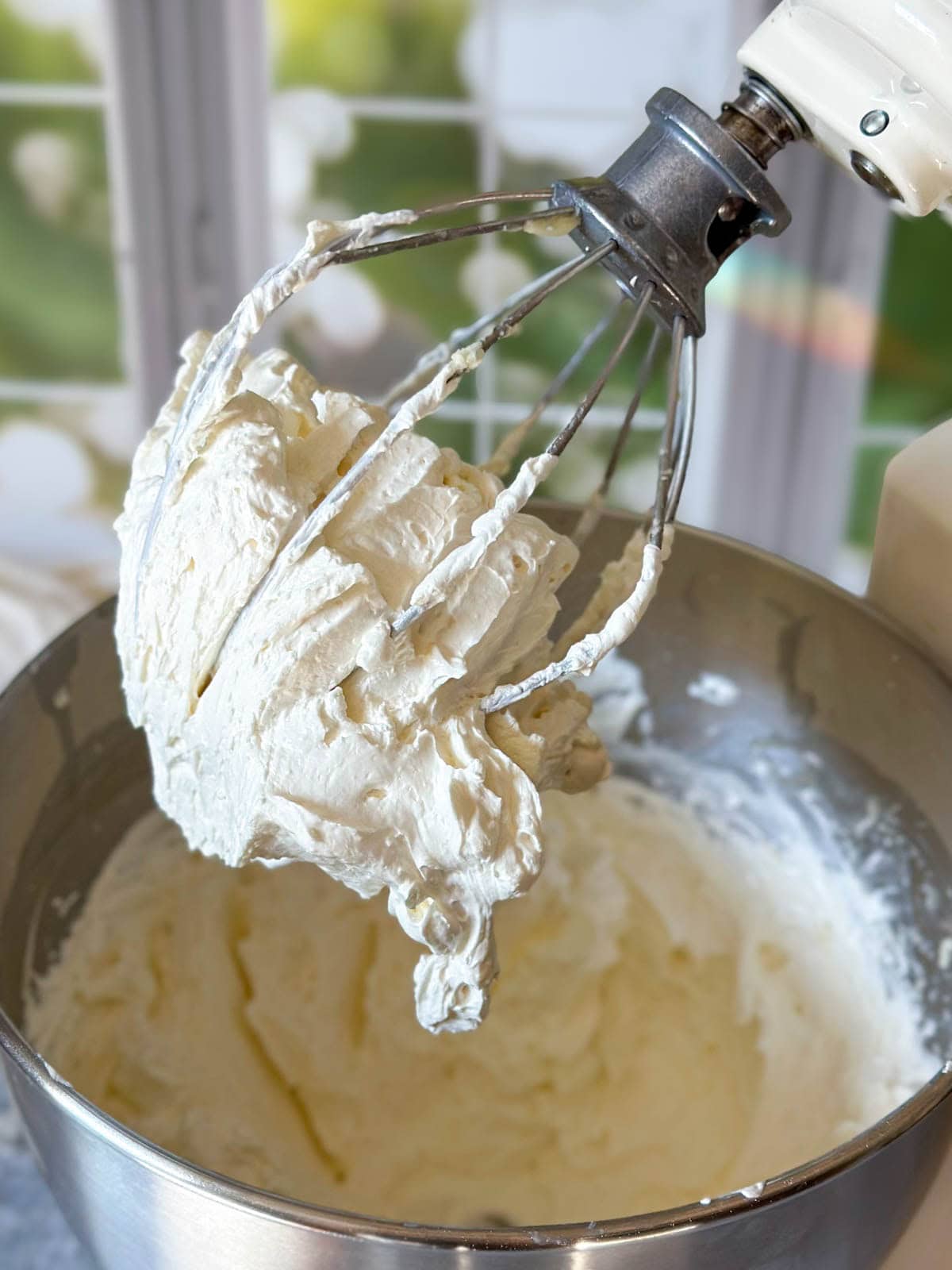 A close up of a stand mixer with the whisk head raised and full of light, fluffy, creamy white whipped body butter