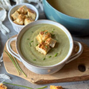 A bowl of creamy broccoli soup with a few croutons on top for garnish. Beyond is a small ramekin of croutons and a large pot full of the remainder of the soup.