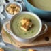 A bowl of creamy broccoli soup with a few croutons on top for garnish. Beyond is a small ramekin of croutons and a large pot full of the remainder of the soup.