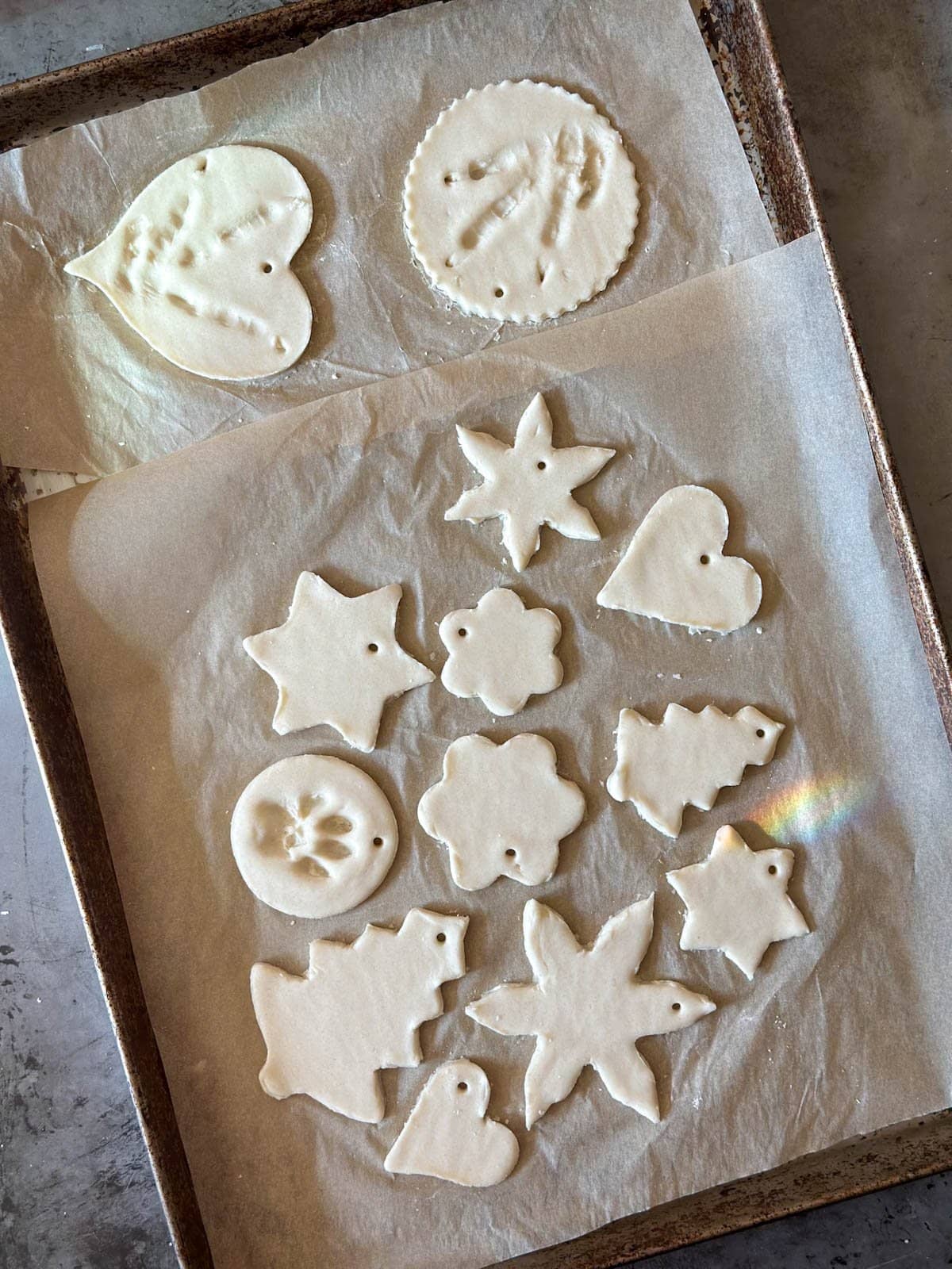 Raw salt dough ornaments i various shapes are spread out on parchment paper on top of a baking sheet.