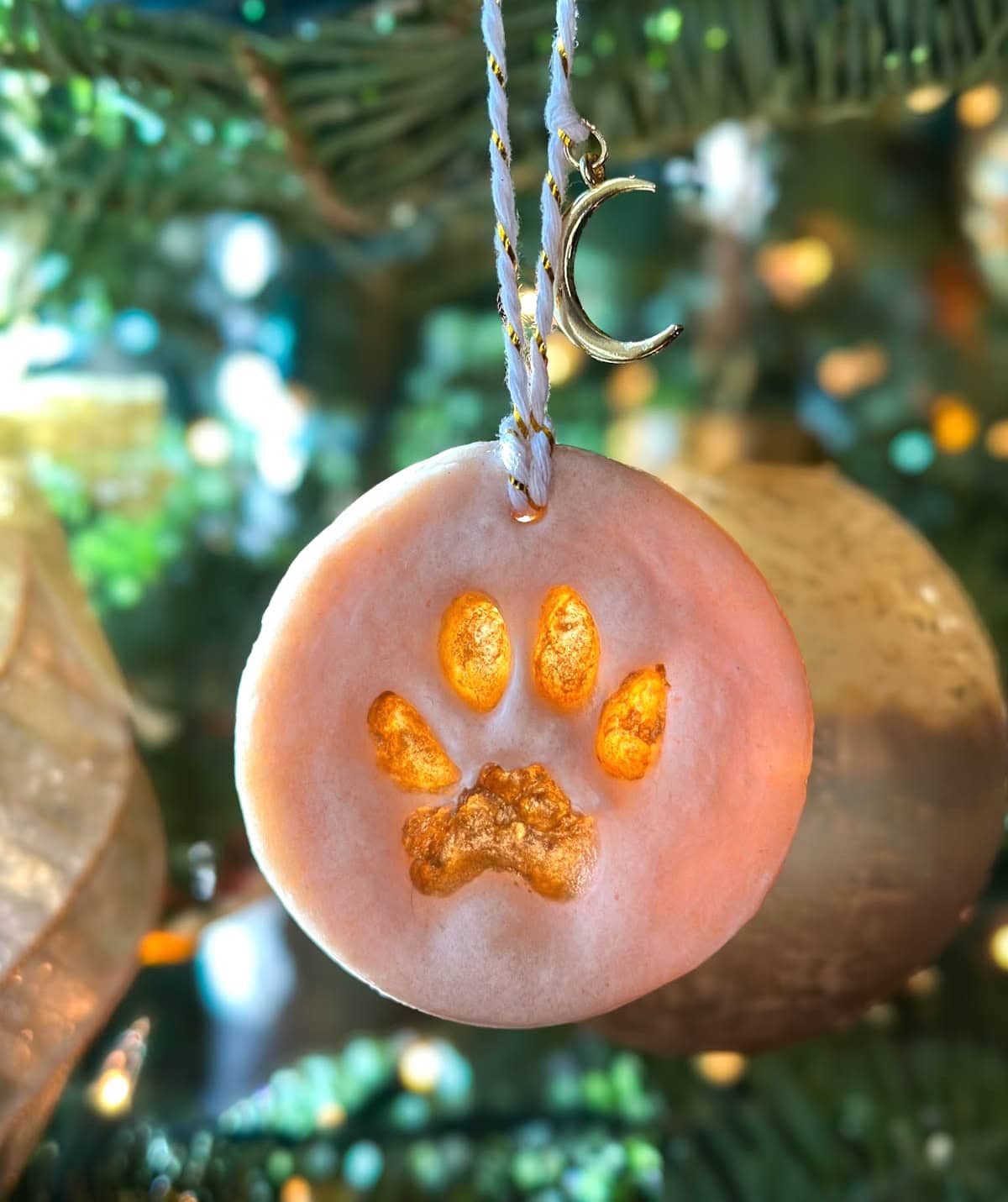 A salt dough ornament with a paw print, white ribbon, and a golden crescent moon attached to the ribbon above the ornament.