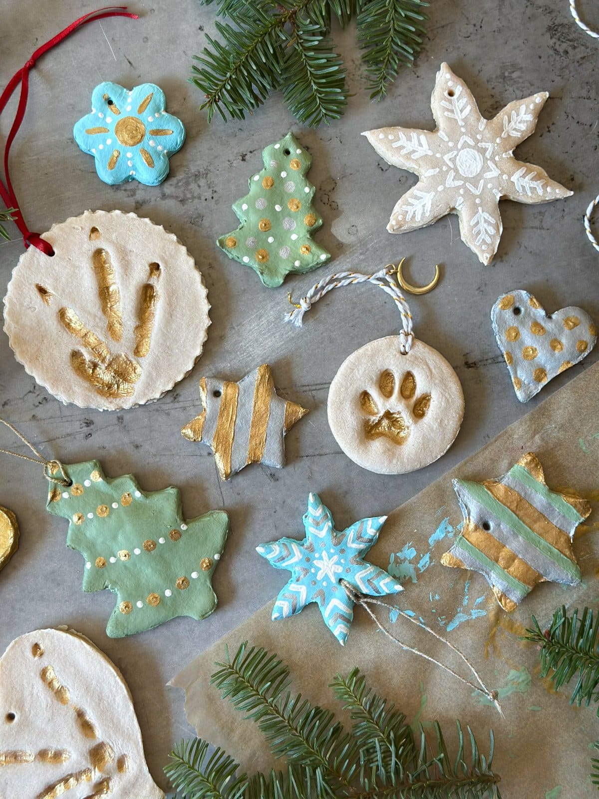 Salt dough ornaments in various shapes and sizes, some fully painted and others only partially are arranged amongst one another. A few pine sprigs adorn the area around the ornaments.