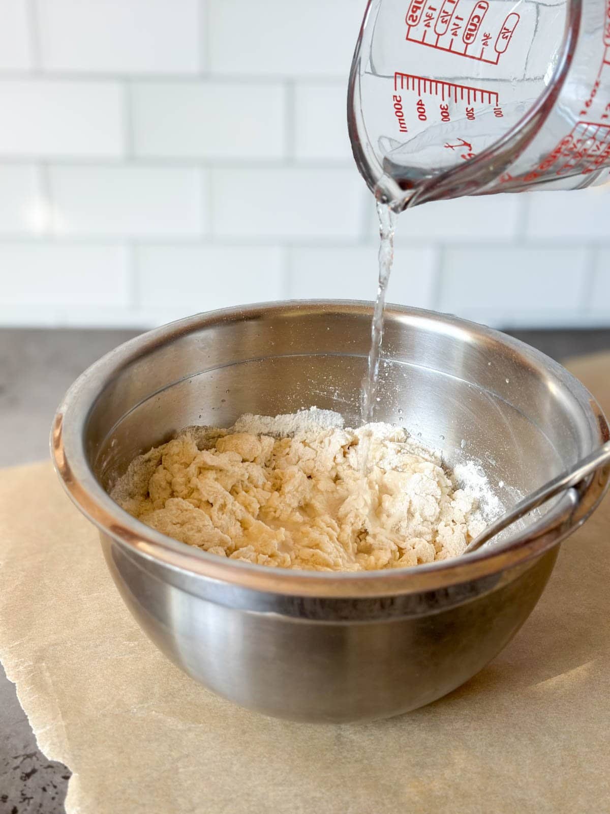 A glass liquid measuring cup held above a metal bowl while water is pouring into the bowl of flour and salt.