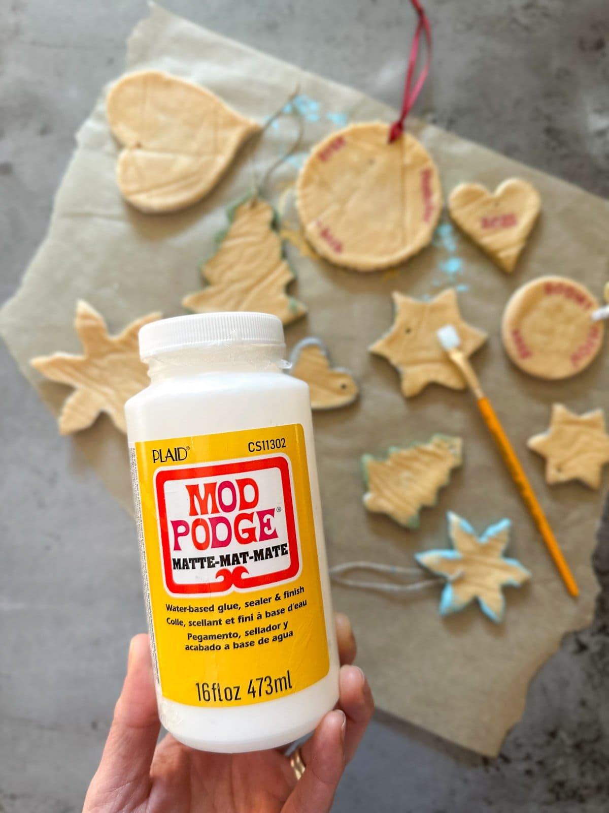 A hand holding a bottle of Mod Podge with ornaments in a variety of shapes in the background.