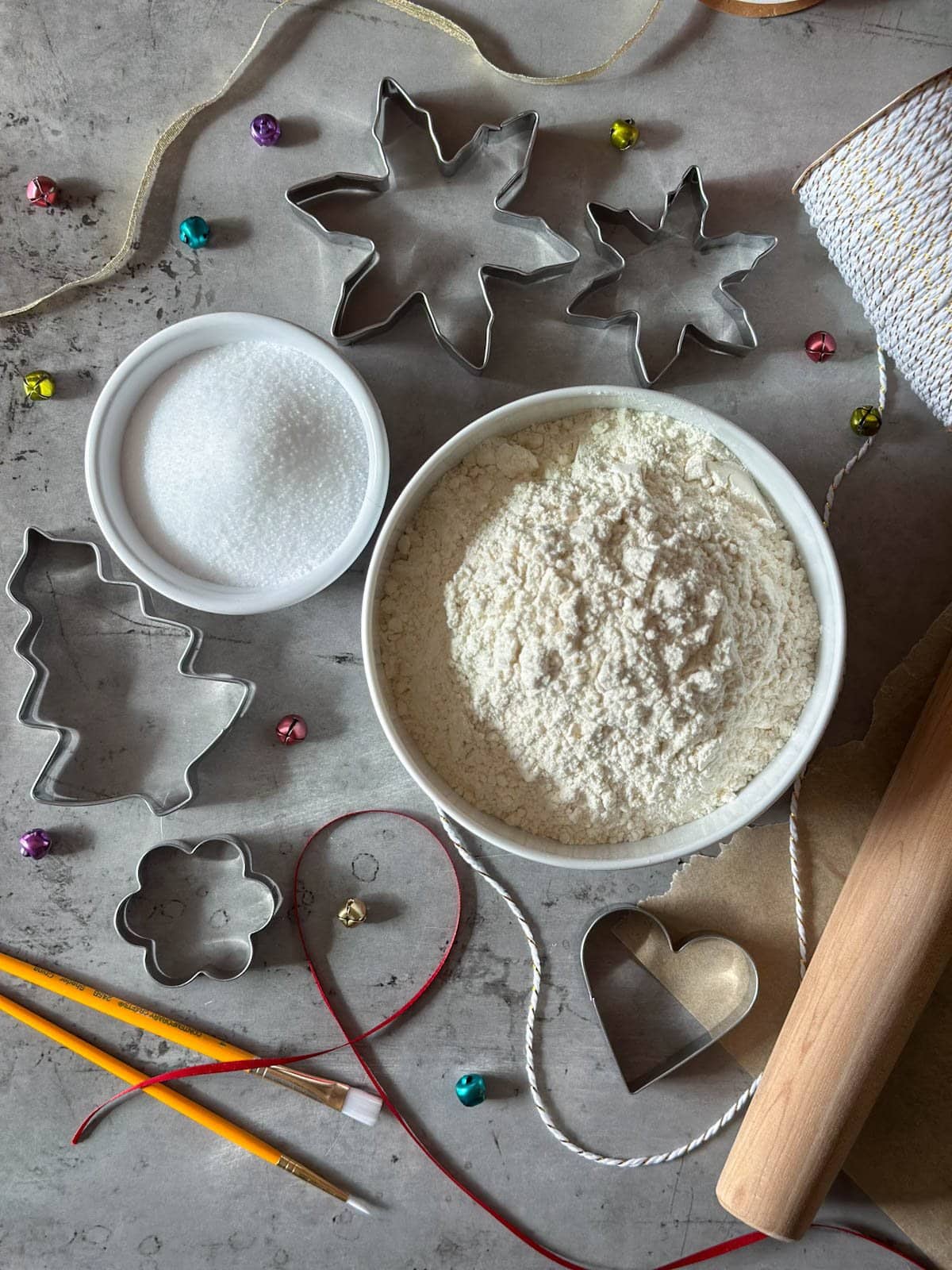 A bowl of flour next to a smaller bowl of salt surrounded by a variety of cookie cutters, ribbon, bells, paint brushes, and rolling pin.