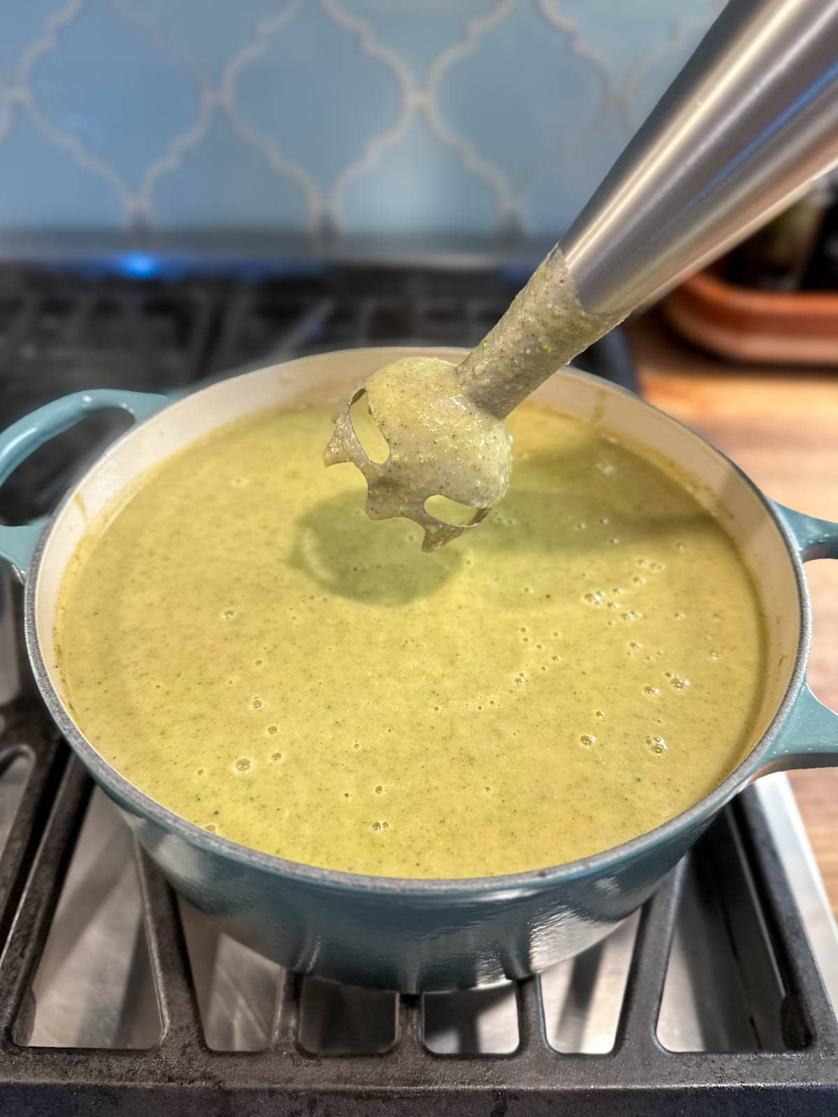 A pot of creamy broccoli soup after it has been blended by an immersion blender which is just being removed from the pot. 