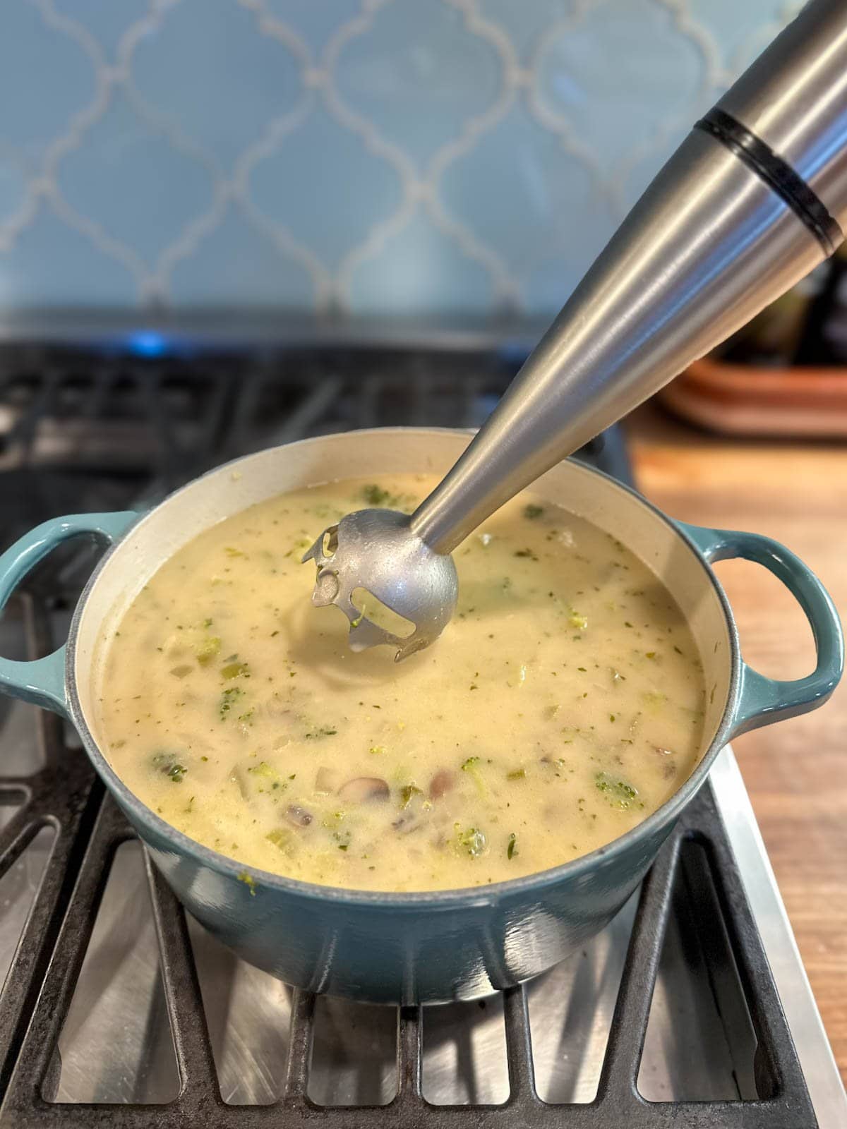 A pot of creamy broccoli soup before it is fully blended. An immersion blender is held above the pot of soup before blending. 