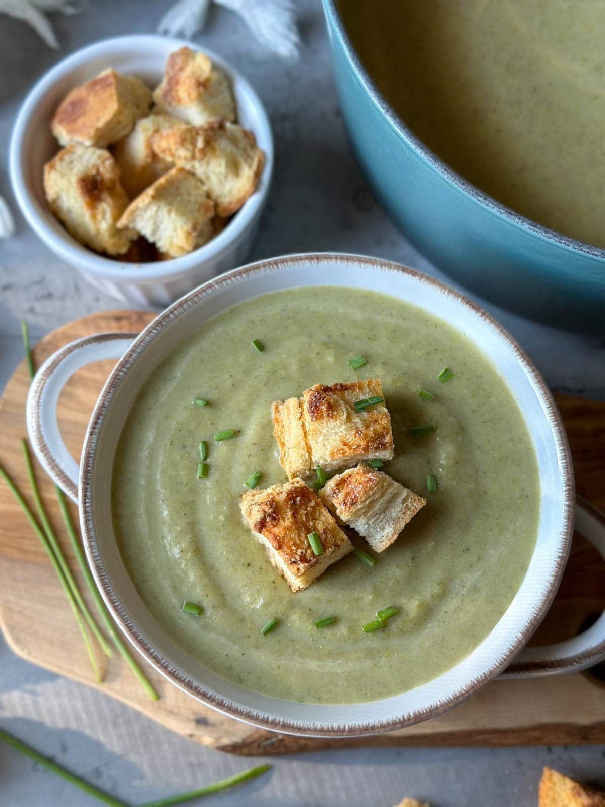 A bowl of creamy broccoli soup garnished with croutons sits next to a small ramekin of croutons and a large pot of the remaining soup. 