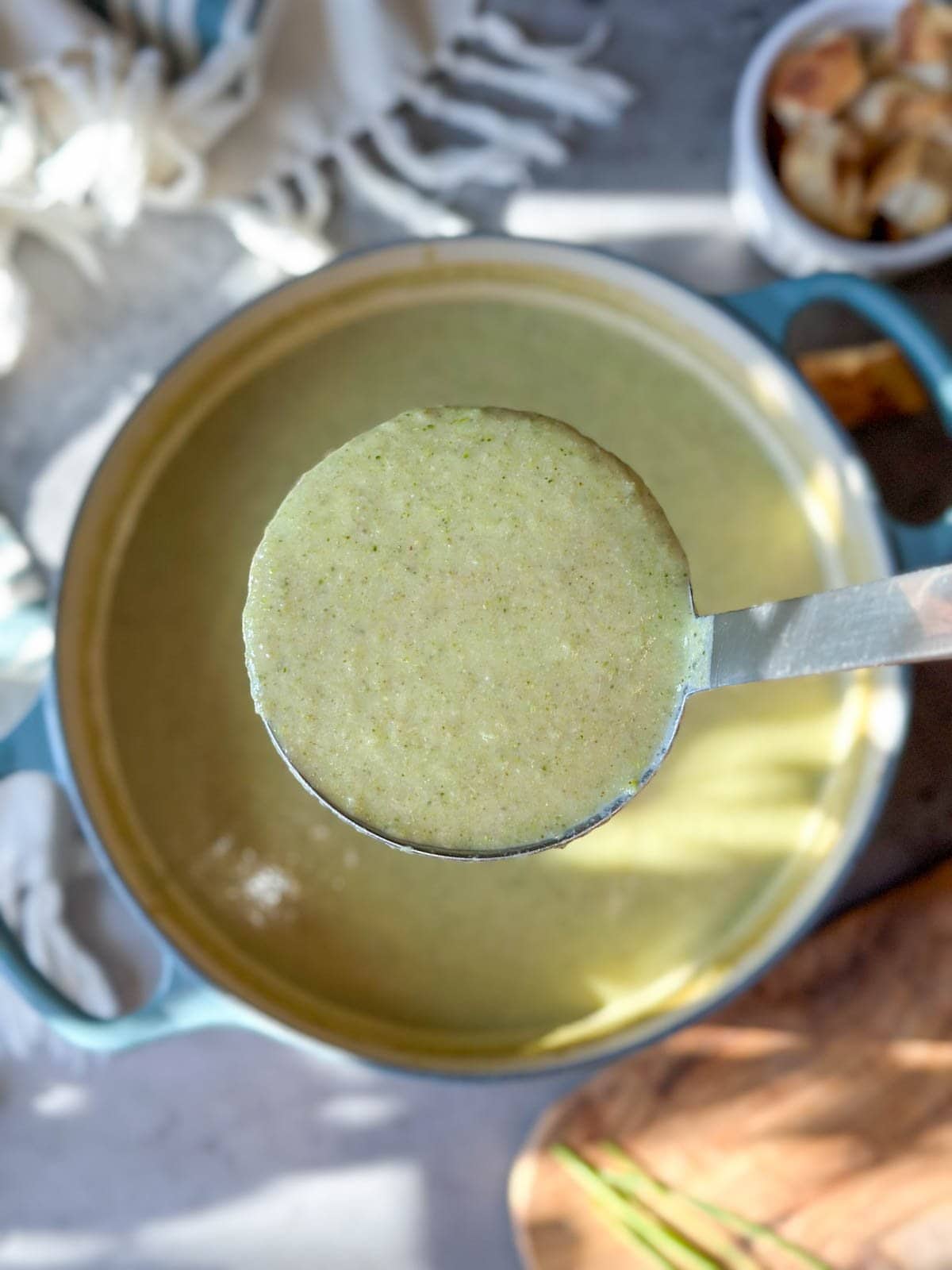 A ladle full of creamy broccoli soup is held above a pot of the same soup. 