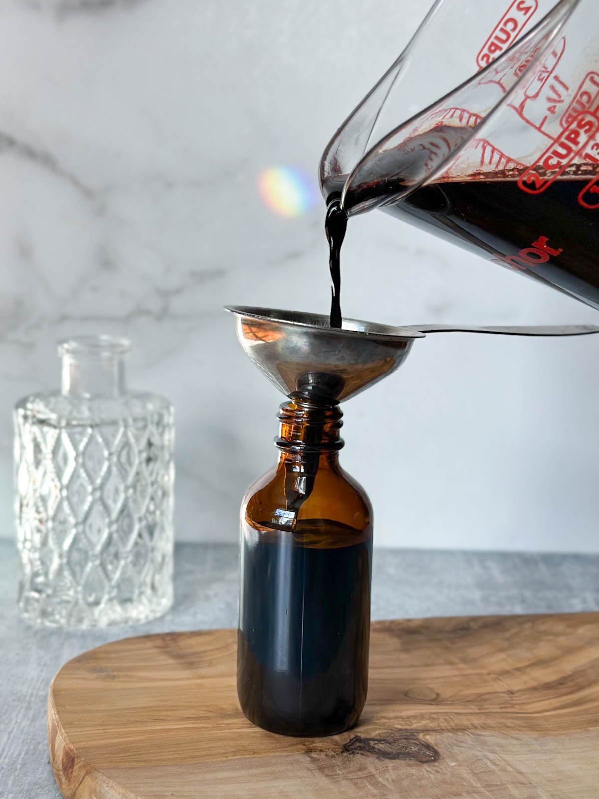 A liquid glass measuring cup is pouring homemade elderberry tincture through a small metal funnel into an amber dropper bottle.
