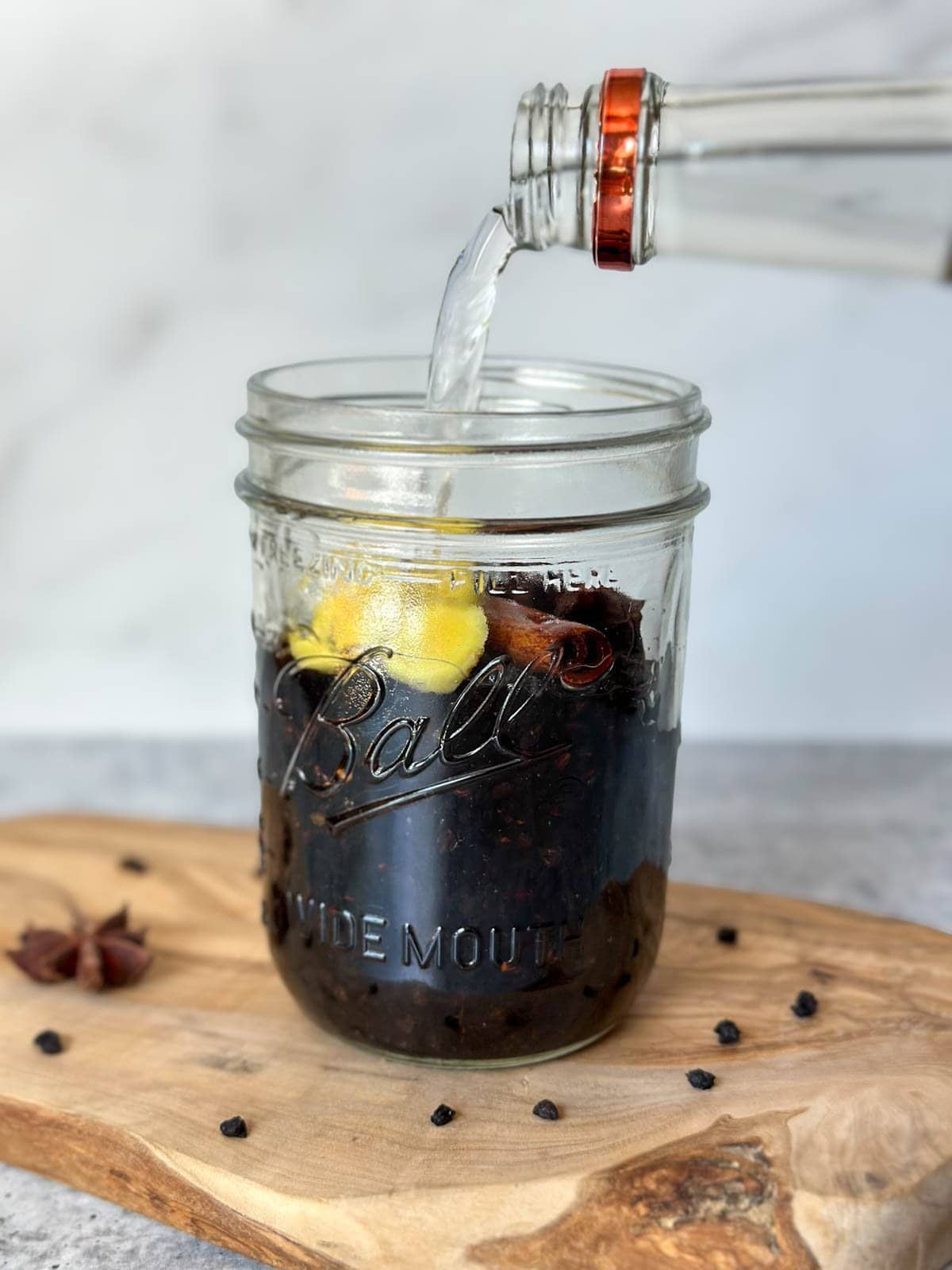 A bottle of clear alcohol is being poured into a pint mason jar of dried berries, cinnamon stick, star anise, and fresh ginger slices.