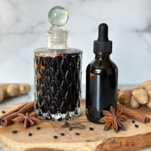 An amber glass dropper bottle sits next to a quilted glass jar, both contain homemade elderberry tincture. Dried elderberries, cinnamon sticks, star anise, and fresh ginger adorn the surrounding area.