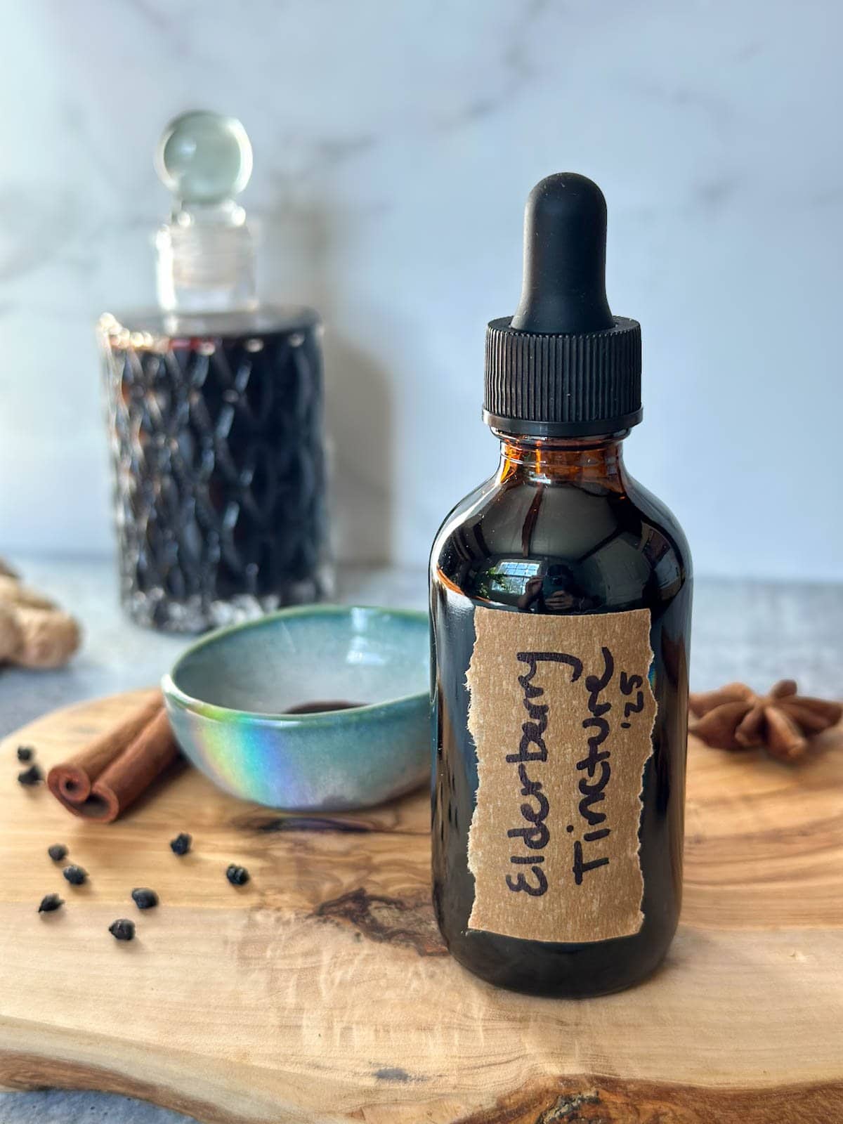 A small amber dropper bottle full of homemade elderberry tincture, dried cinnamon sticks, star anise, ginger, and elderberries adorn the surrounding area. Beyond is a quilted glass jar filled with the remaining elderberry tincture.