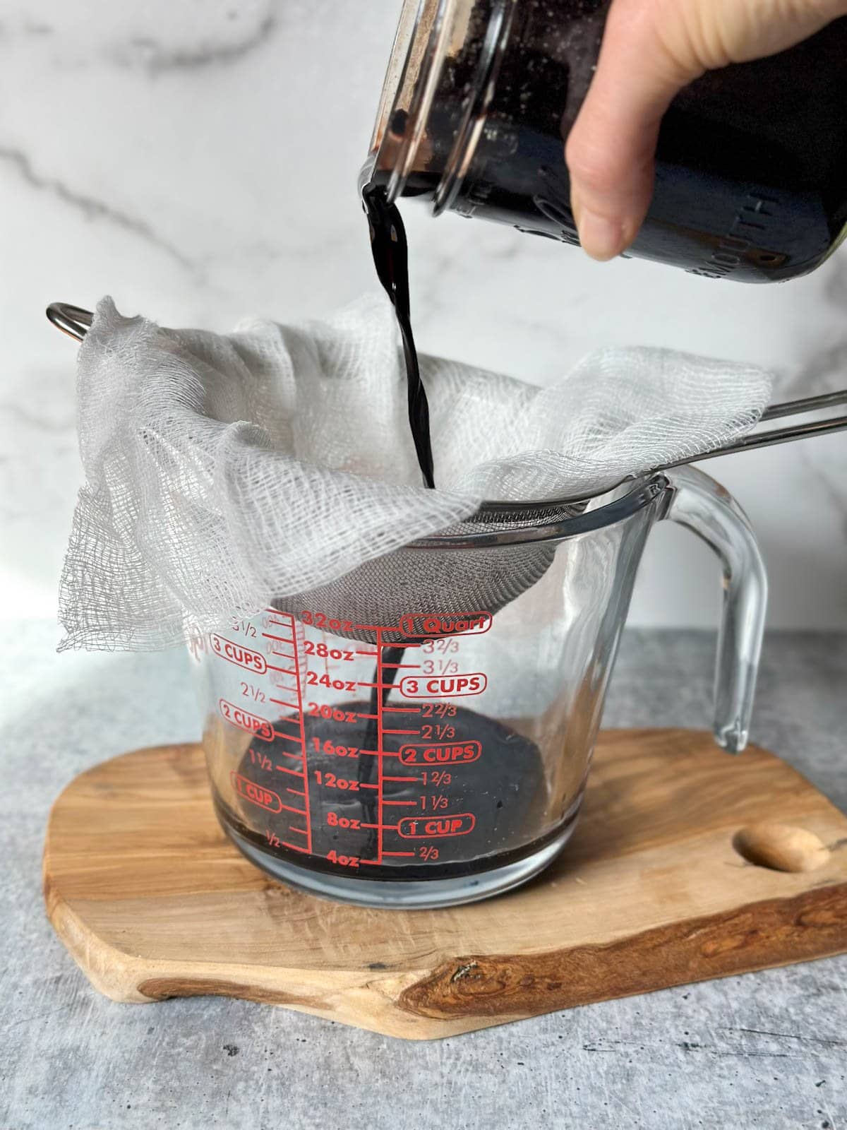 Cheesecloth lines a fine mesh strainer which sits on top of a liquid glass measuring cup. Homemade elderberry tincture is being poured into the strainer.
