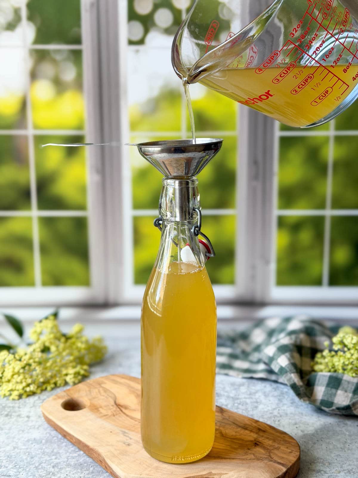 A liquid glass measuring cup is pouring elderflower syrup into a large bottle with a flip top lid using as stainless steel funnel. There are a few bunches of elderflowers arranged nearby. 
