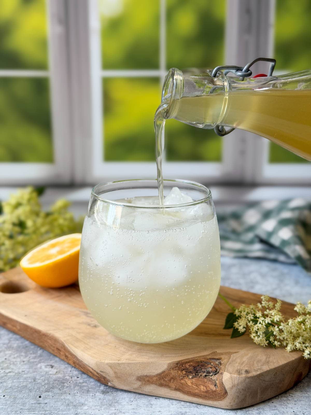 A glass bottle with a flip top lid is pouring liquid into a stemless wine glass that is almost full of carbonated liquid along with several ice cubes. 