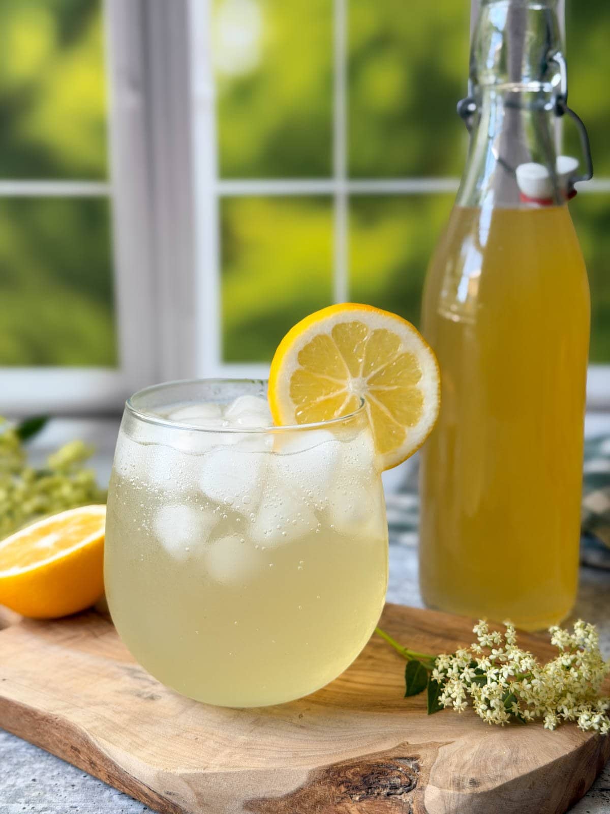 A stemless wine glass with elderflower cordial and sparkling water with a lemon wedge on the edge of the glass. A large bottle of elderflower cordial recipe is nearby along with flowers and lemon cut in half. 