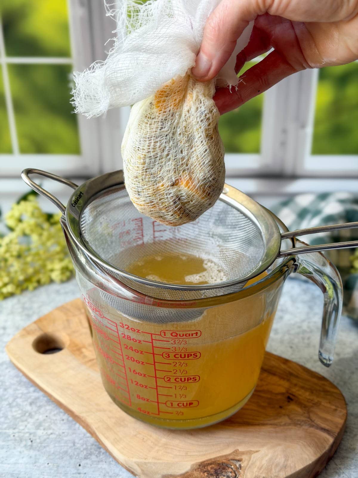A fine mesh strainer is sitting on top of a glass liquid measuring cup full of elderflower cordial syrup. A wad of cheesecloth is being held above the strainer which has been used to further strain the infused contents while being able to wring out the juices. 