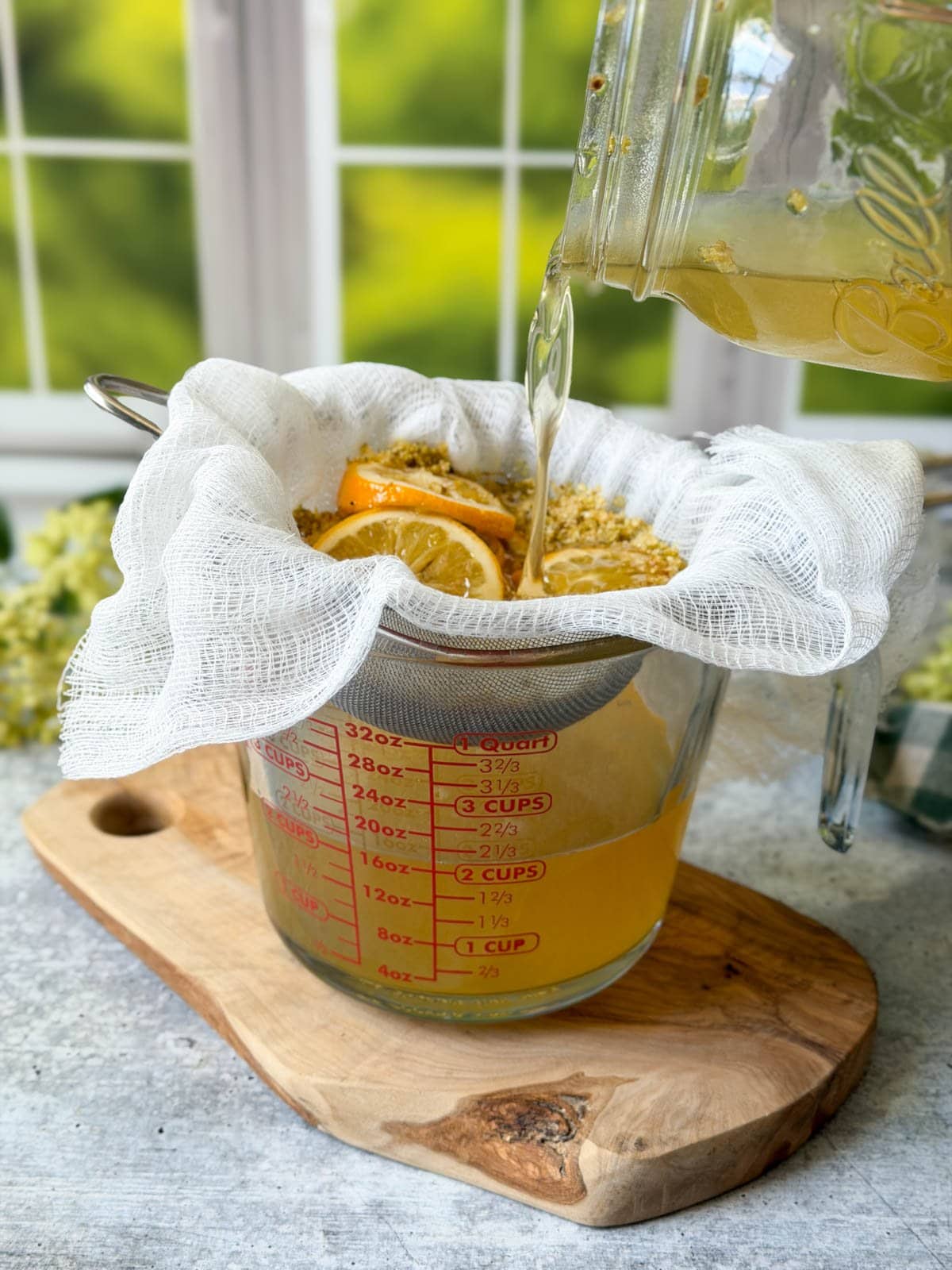 A mason jar is pouring elderflower cordial syrup through a section of cheesecloth sitting on top of a fine mesh strainer into a liquid glass measuring cup. 