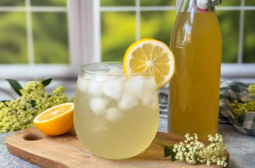 A stemless wine glass of sparkling water mixed with elderflower cordial syrup with a lemon wedge on the edge of the glass. The bottle of elderflower cordial syrup sits nearby along with fresh elderflower blossoms and half a lemon.