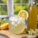 A stemless wine glass of sparkling water mixed with elderflower cordial syrup with a lemon wedge on the edge of the glass. The bottle of elderflower cordial syrup sits nearby along with fresh elderflower blossoms and half a lemon.