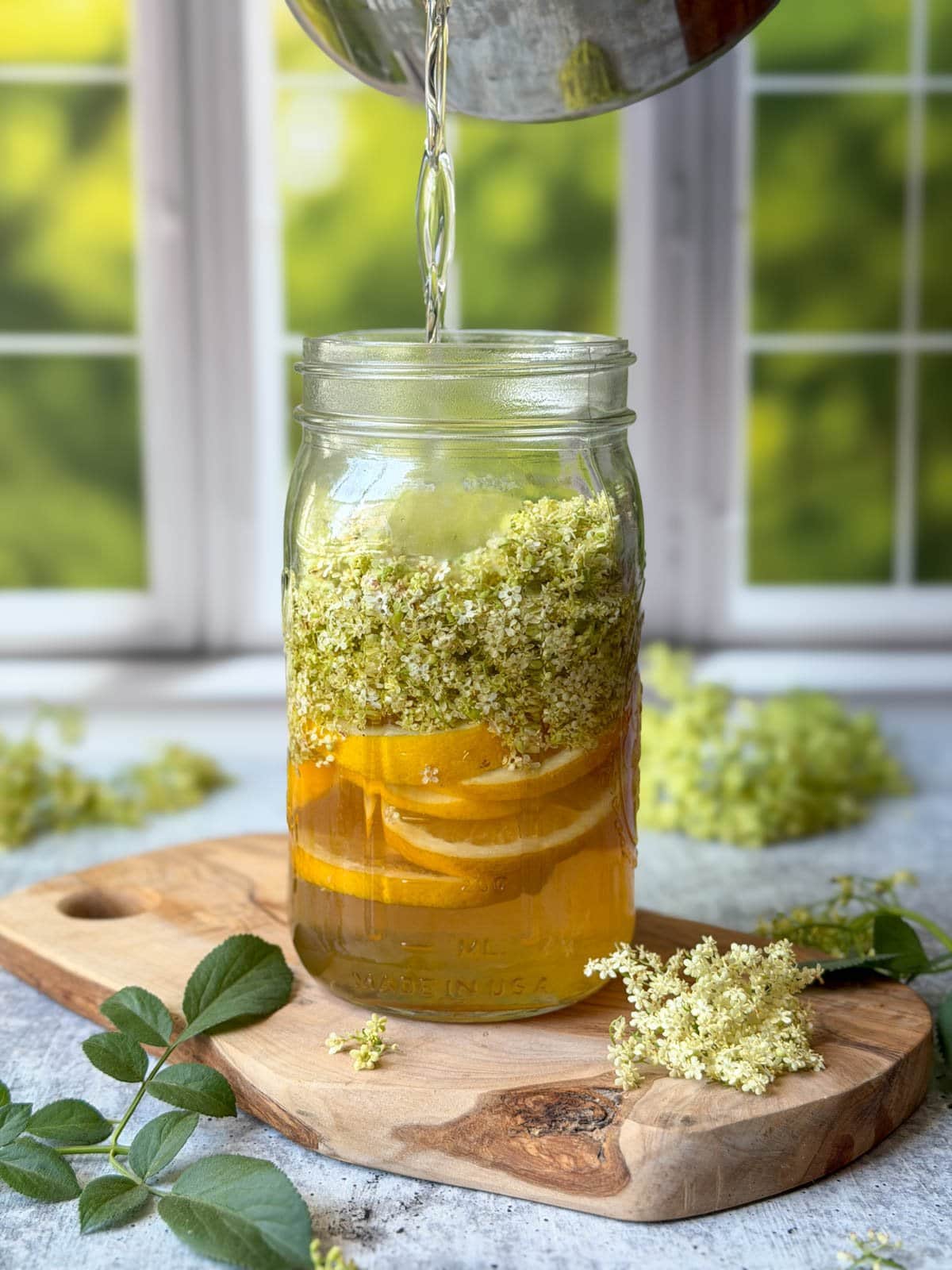 A simple syrup solution is being poured from a pot into a quart mason jar full of slices of lemons and flower blooms. 