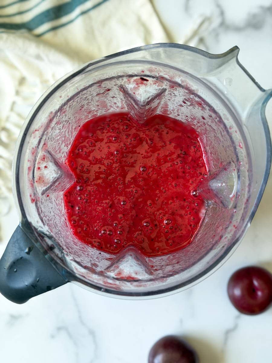 Freshly blended fruit puree inside of a blender. A fresh plum sits nearby. 