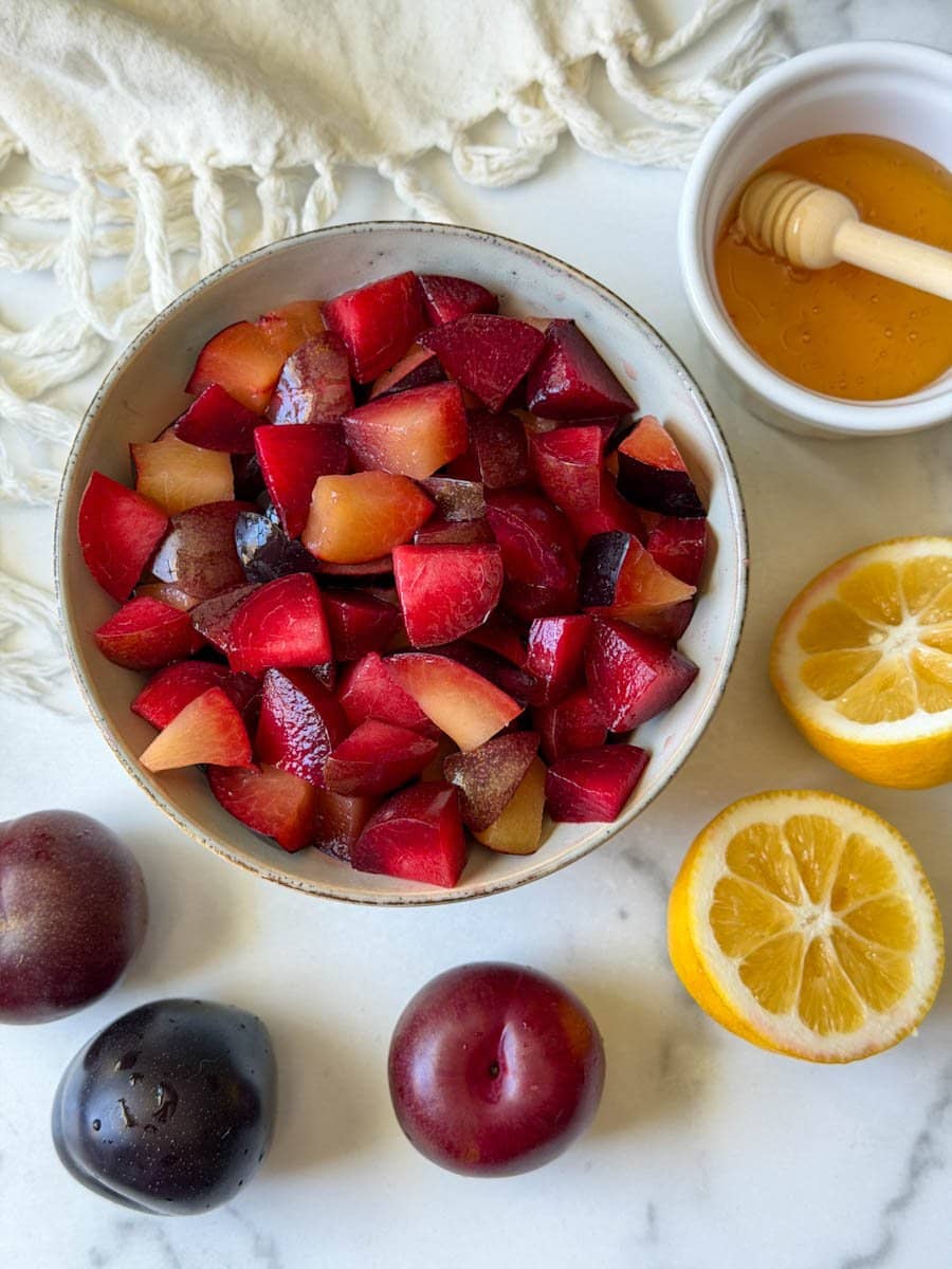 A ceramic bowl is full of chopped plums while a small ramekin nearby is partially full of honey, while a fresh lemon cut in half and whole plums sit nearby. 