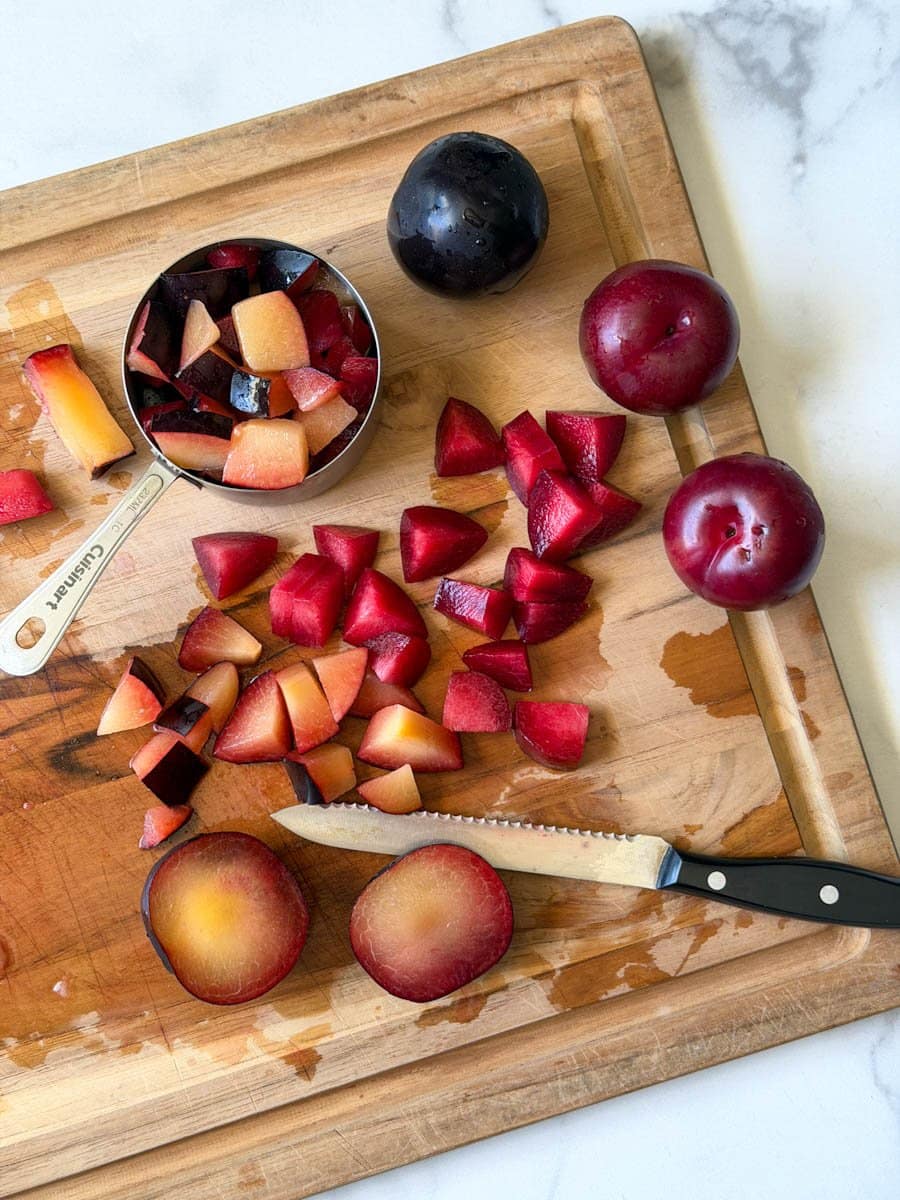 Fresh plums are being chopped into pieces on a wood cutting board. A metal measuring cup is full of some of the chopped plums while many pieces and a few whole plums remain on the board. 