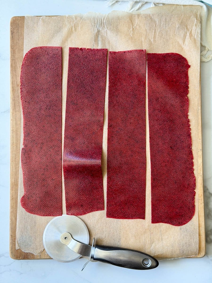 A piece of parchment paper rolled out onto a wood cutting board has strips of fruit leather which have been cut with a pizza slicer. 
